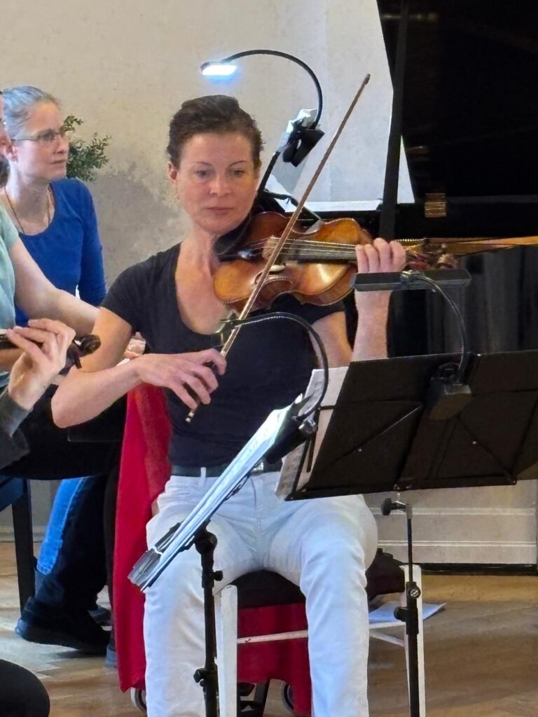 Frau in dunkelgrauem Oberteil und weißer Hose spielt Violine, vor ihr ein beleuchteter Notenständer. Links teilweise eine weitere Violinistin sichtbar, im Hintergrund sitzt eine Frau im blauen Oberteil am Klavier. Innenraum mit Holzfußboden und Pflanze.