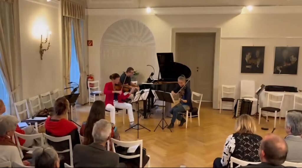 Drei Musiker:innen spielen klassische Kammermusik in einem hellen Raum mit Holzfußboden und weißer Wand. Publikum sitzt auf weißen Stühlen und hört aufmerksam zu. Ein Akkordeon steht rechts auf einem Stuhl.
