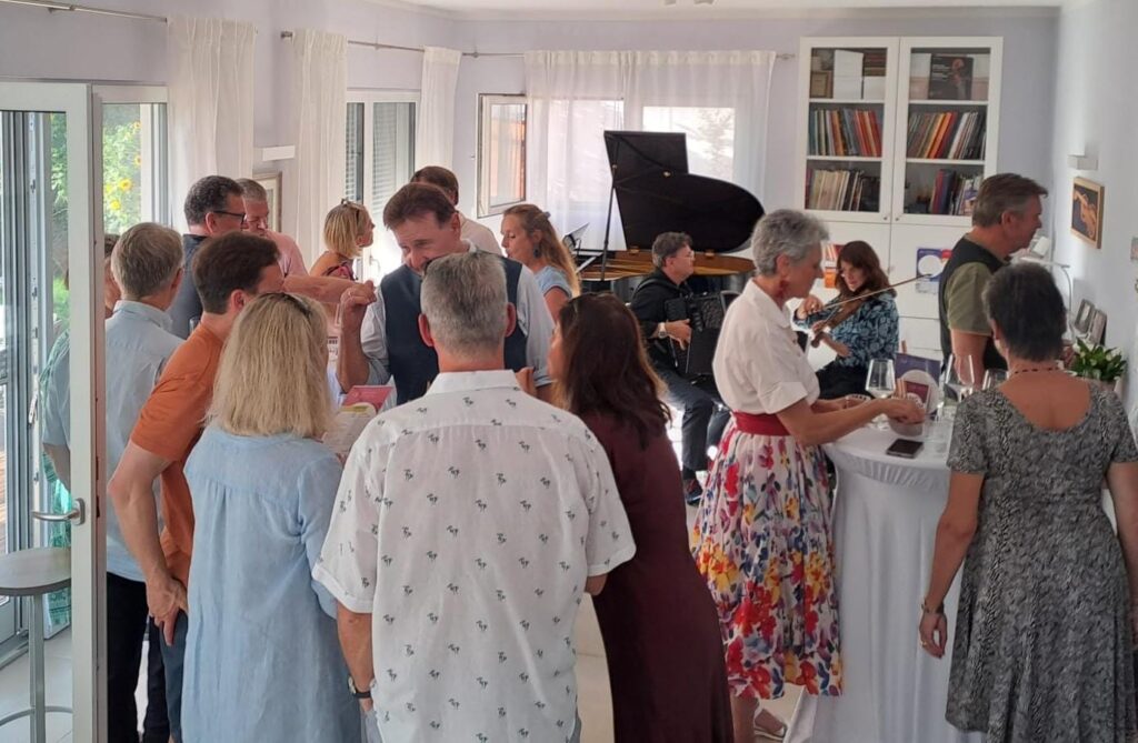 Gesellige Runde von etwa fünfzehn Personen in hellem Raum mit großen Fenstern; im Hintergrund Violinist und Flügel, Bücherregal und Schallplatten; entspannte Atmosphäre