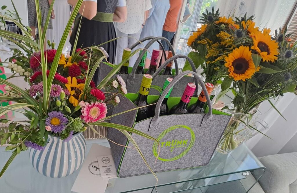 Zwei graue Filztaschen mit grünem TRUNK-Logo und Flaschen vom Weingut TRUNK, umgeben von Blumensträußen (Streifen-Vase, Sonnenblumen) auf einem Tisch; Menschen im Gespräch im Hintergrund