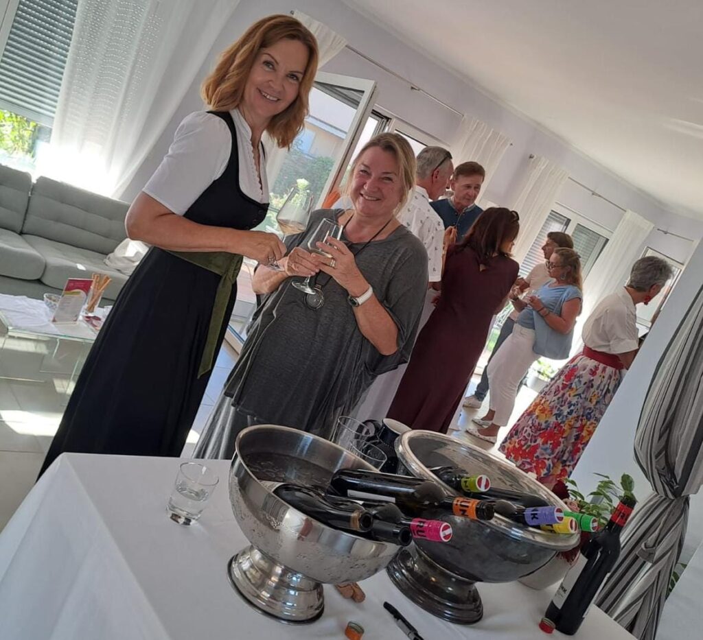 Zwei lächelnde Frauen mit Weingläsern neben Tisch mit zwei Metallschüsseln voller Weinflaschen; gesellige Feier in modernem, hellen Raum