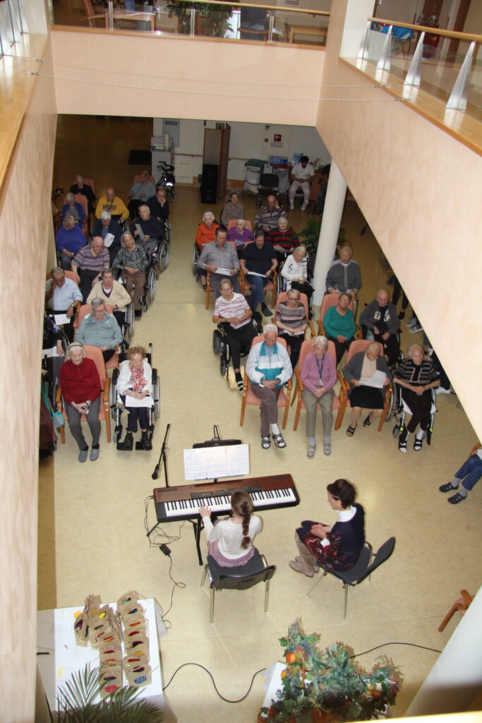 Musikalische Darbietung im Seniorenheim: Ein Mädchen spielt E-Piano, eine Frau steht daneben, während ältere Menschen – einige in Rollstühlen – in einem hellen, freundlich dekorierten Gemeinschaftsraum aufmerksam zuhören.