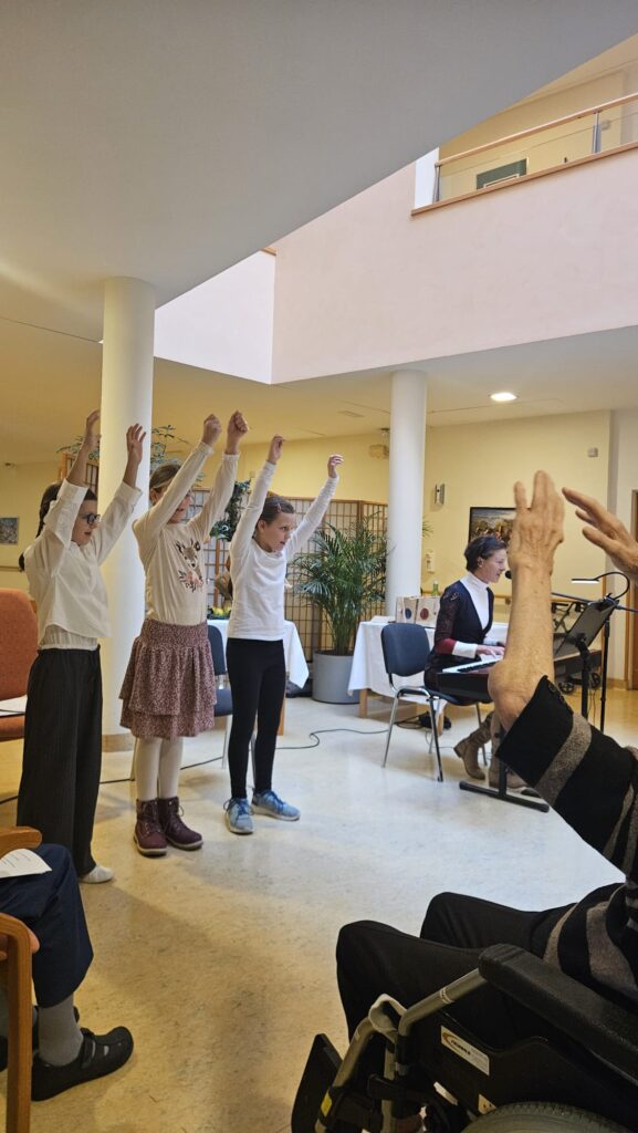 Kinder führen eine musikalische Tanzaufführung in einem Seniorenzentrum auf, begleitet von Pianomusik, vor einem Publikum mit älteren Menschen, darunter eine Person im Rollstuhl
