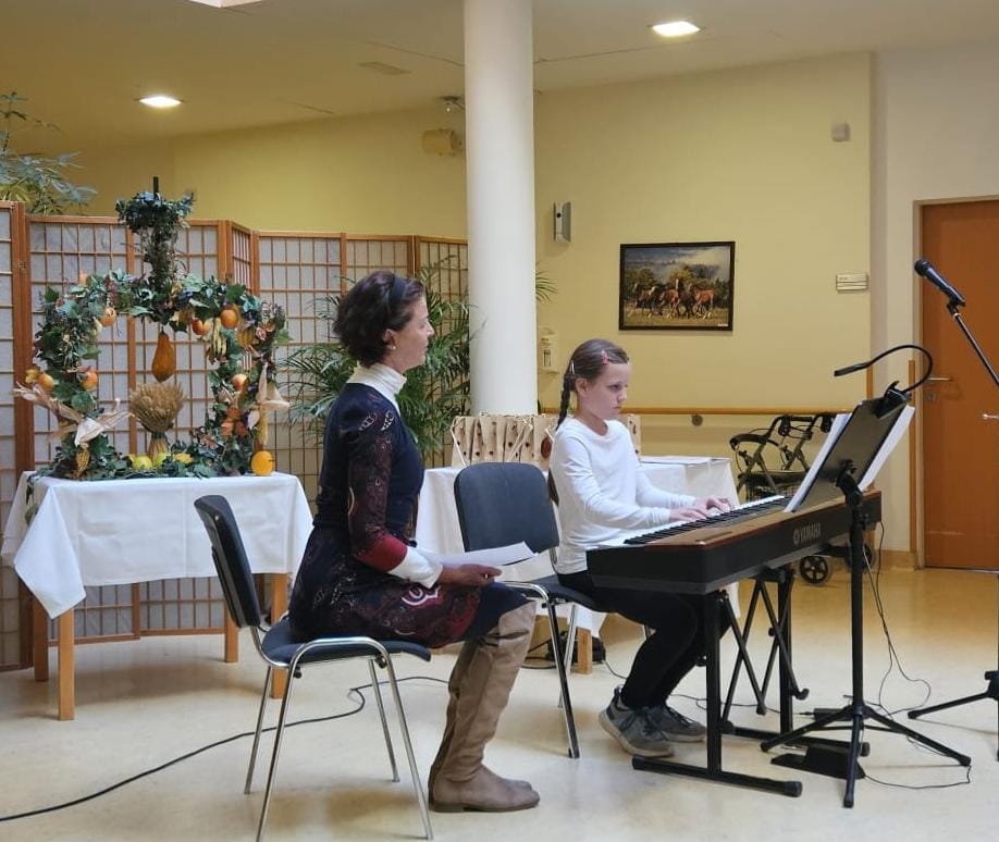 Zwei Personen bei herbstlich dekorierter Musikaufführung – eine spielt Klavier, die andere hält Notenblätter.
Im Hintergrund herbstliche Dekoration mit Kürbissen und Laub.