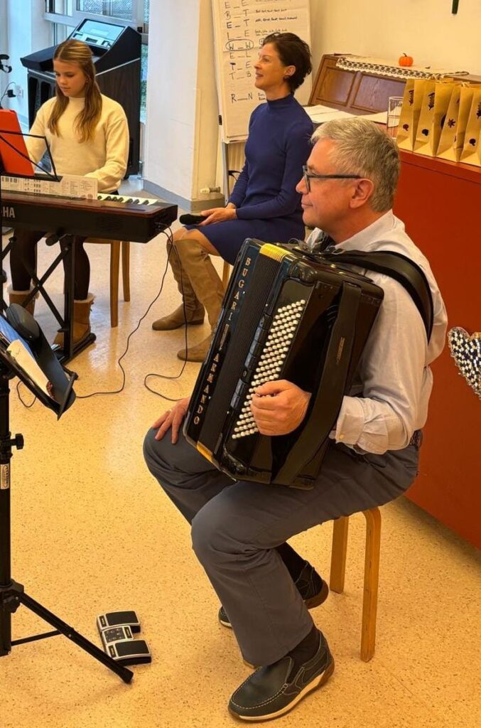 Drei Personen musizieren in der Aula eines Altenheims: Eine Jugendliche spielt E-Piano, eine Frau hält ein Mikrofon, ein Mann spielt Akkordeon.