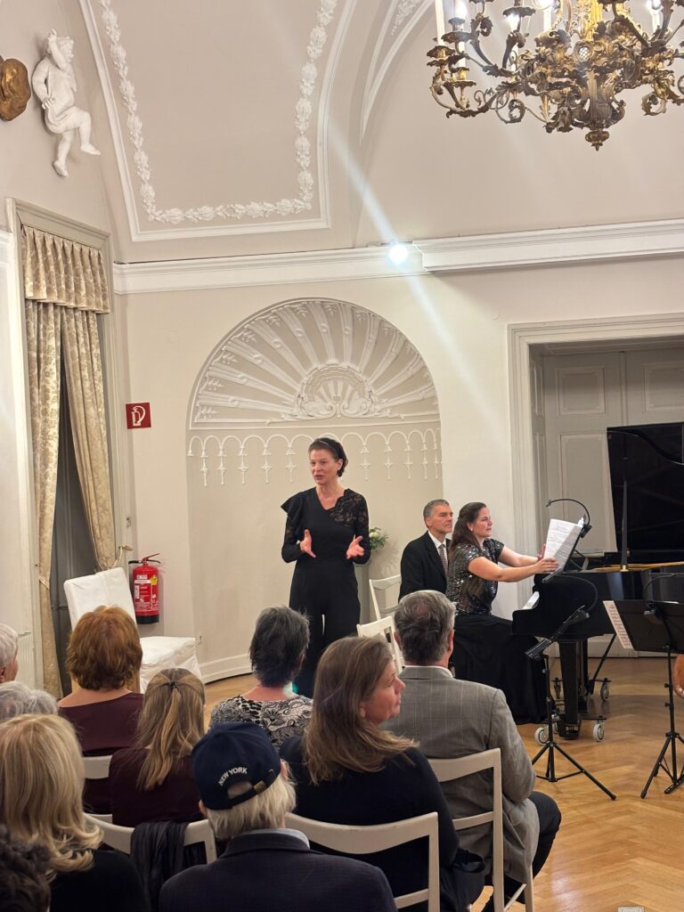 Moderation einer musikalischen Veranstaltung im Festsaal des Thonetschlössls in Mödling: Eine Frau in schwarzer Kleidung spricht vor Publikum, neben ihr ein Pianist am Flügel. Der prunkvolle Saal ist mit Stuckdecke, Kronleuchter und Wandskulptur ausgestattet.