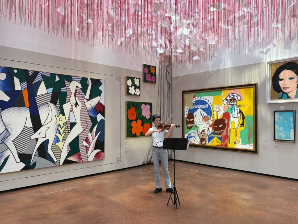 Eine Musikerin spielt Violine in einem Ausstellungsraum mit farbenfrohen, modernen Kunstwerken und hängenden rosa‑weißen Papierstreifen an der Decke. Die Verbindung von Musik und bildender Kunst schafft eine lebendige Atmosphäre während des Rundgangs „Musik geht mit Kunst!“.