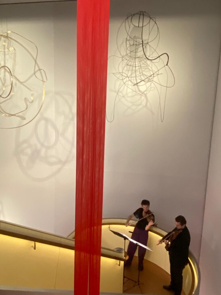 Zwei Musikerinnen spielen Streichinstrumente am Fuß einer spiralförmigen Rampe mit gelb beleuchtetem Handlauf. An der Wand hängen abstrakte Drahtskulpturen, im Zentrum hängt eine vertikale rote Stoffinstallation. Die musikalische Intervention begleitet die Ausstellung „Die Linie“ in der Heidi Horten Collection.