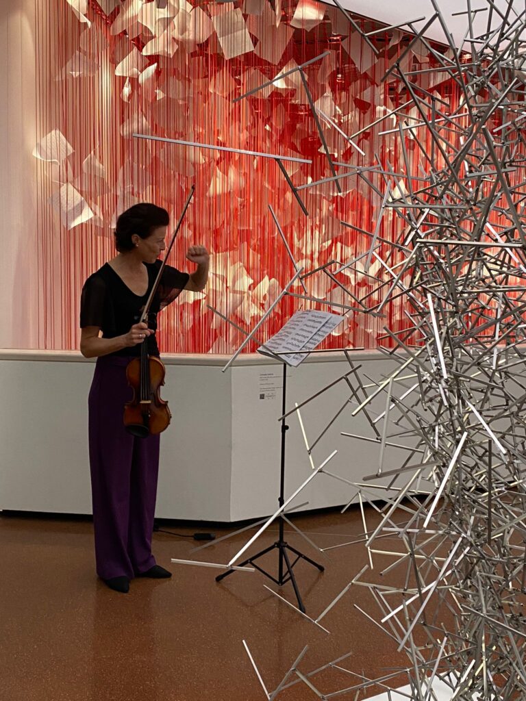 Eine Musikerin mit Violine steht in einer Galerie vor einer Installation aus roten, vertikal gespannten Fäden und schwebenden weißen Papierfragmenten. Vor ihr befindet sich ein Notenständer, daneben eine abstrakte Metallskulptur. Die Szene verbindet Live‑Musik mit zeitgenössischer Kunst im Rahmen der Ausstellung „Die Linie“.
