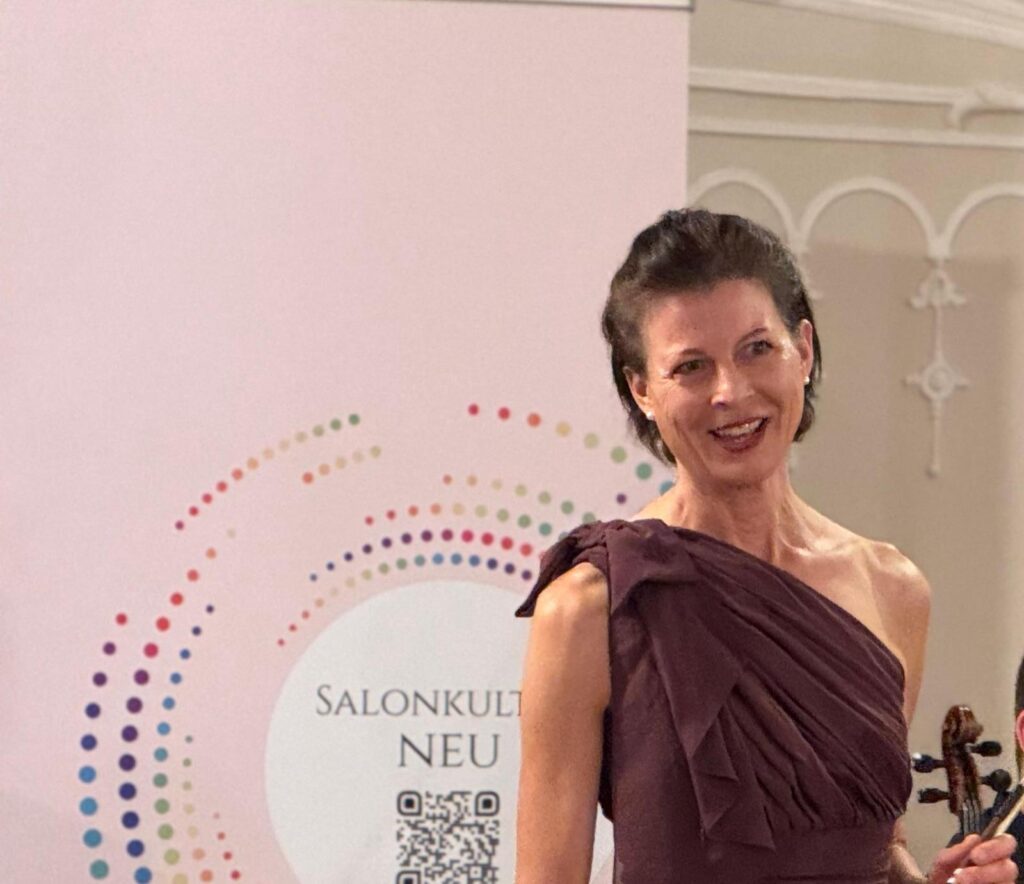 Violinistin in burgunderfarbenem, einseitig schulterfreiem Abendkleid steht vor einem Banner mit der Aufschrift ‚SalonKultur Neu‘ und hält ihre Violine.