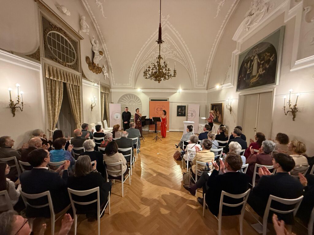 Publikum in einem prunkvollen Saal applaudiert drei Personen auf der Bühne: zwei Musiker in formeller Kleidung und eine Frau im roten Kleid; hinter ihnen stehen Veranstaltungsbanner.