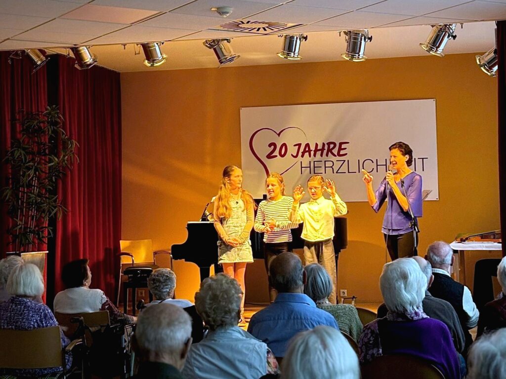 Bühnenauftritt in einem Gemeinschaftsraum: Drei Kinder und eine erwachsene Frau stehen vor einem Publikum älterer Menschen. Die Frau spricht in ein Mikrofon, hinter ihnen stehen ein Flügel und ein Banner mit der Aufschrift ‚20 Jahre Herzlichkeit