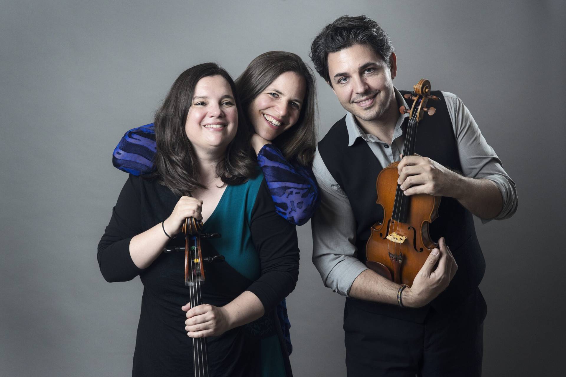 Ein Trio aus drei Musiker*innen des Ensemble Triosonate, eng beieinander vor neutralgrauem Hintergrund stehend; links eine Person mit Cello, rechts eine Person mit Violine, in der Mitte eine Person, die beide mit den Armen umfasst. Die Szene vermittelt Nähe, Zusammenhalt und kammermusikalische Atmosphäre.