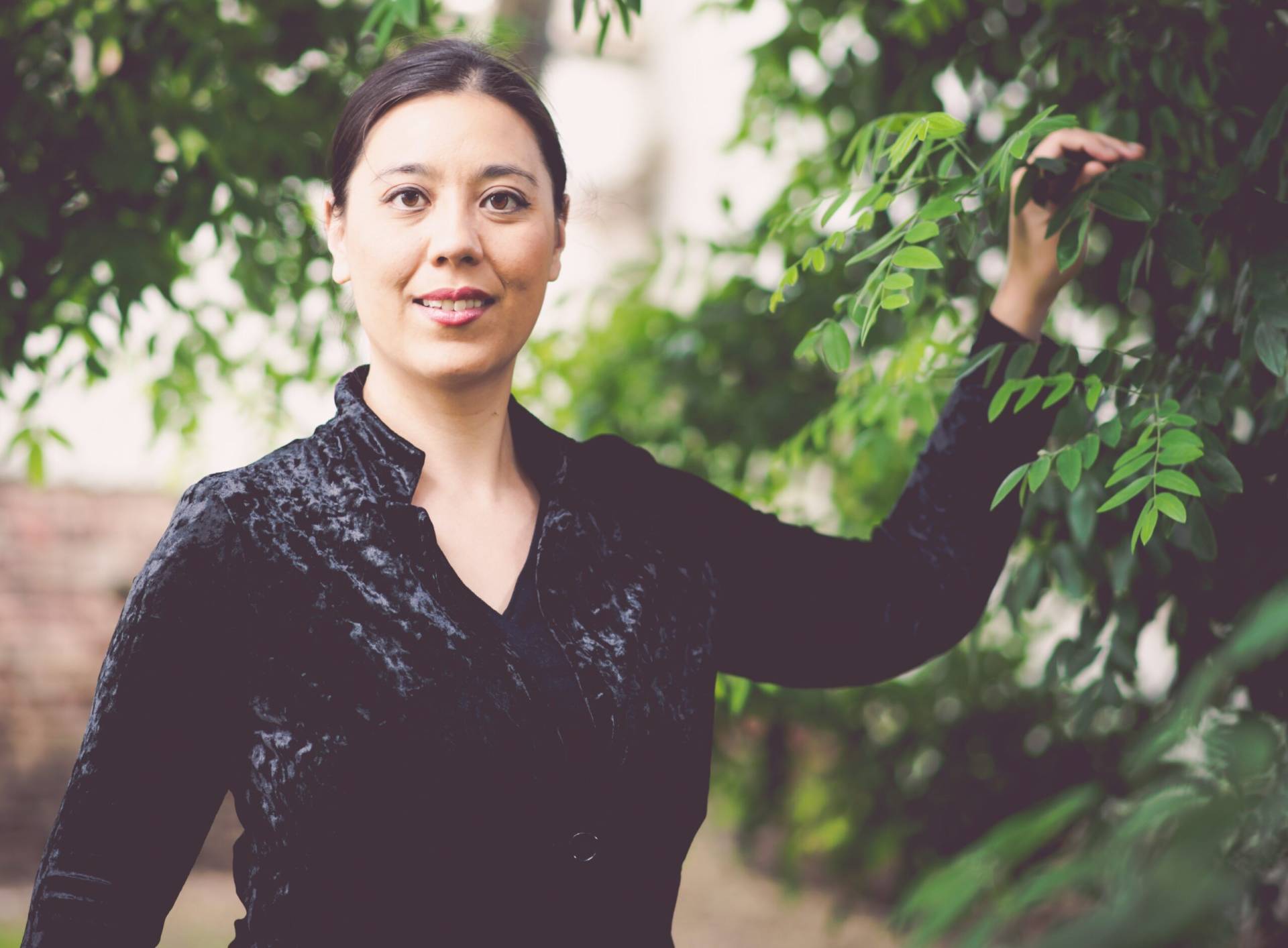 Person (Frau Katharina Gebauer) in schwarzem Oberteil steht draußen und berührt einen grünen Zweig vor natürlichem Hintergrund. Besetzung: Sopran